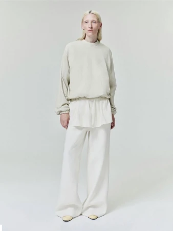 Model wearing neutral toned layered sweatshirt and loose trousers standing against a pale grey seamless studio backdrop