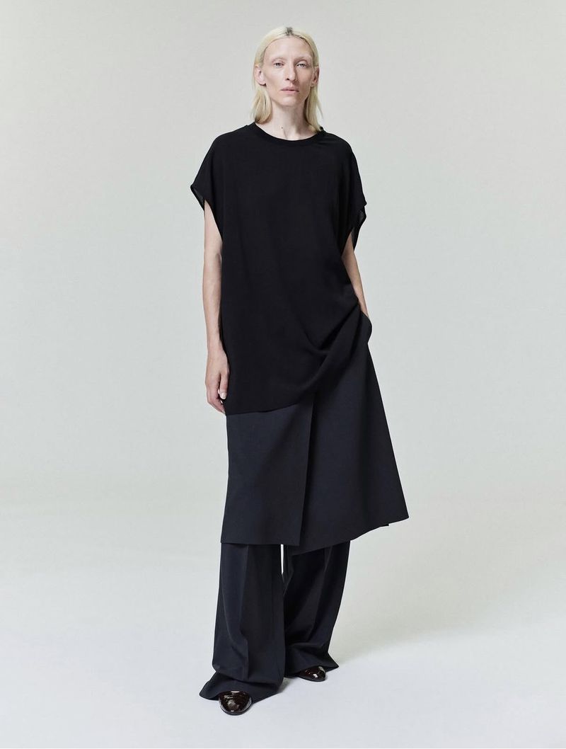 Model standing in a black top and dark loose trousers against a solid grey background for Fear of God.