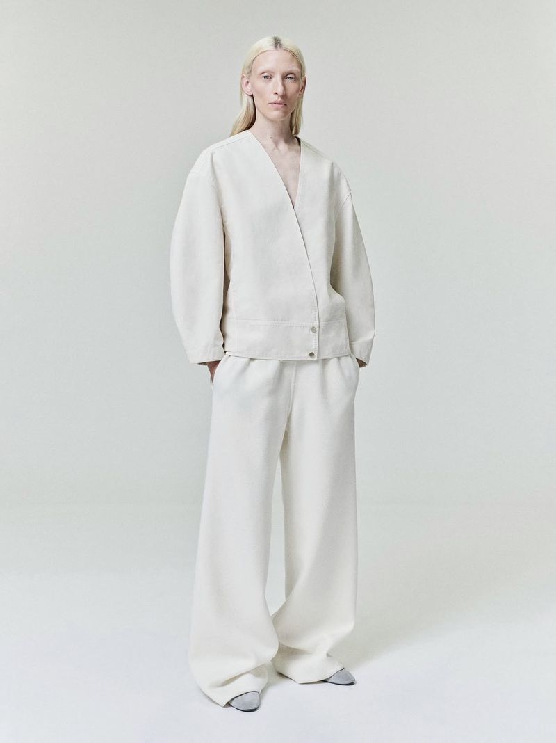 Model standing against a plain white studio background wearing an off-white v-neck jacket and matching wide-leg trousers