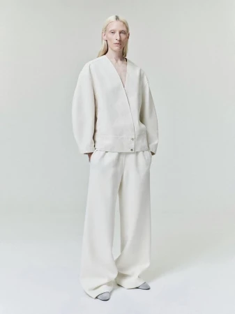 Model standing against a plain white studio background wearing an off-white v-neck jacket and matching wide-leg trousers
