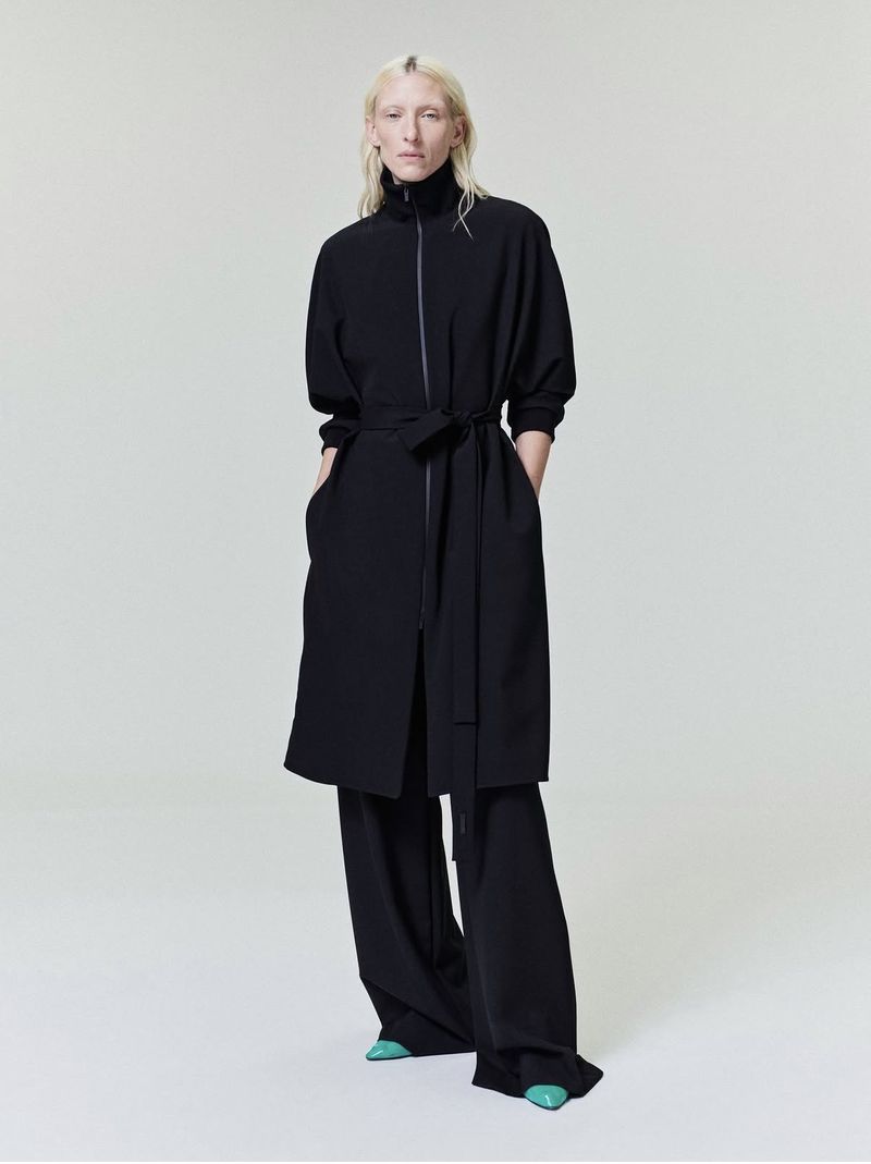 Full-length studio shot of a woman in a black dress with a belt and black pants, standing against a white background.