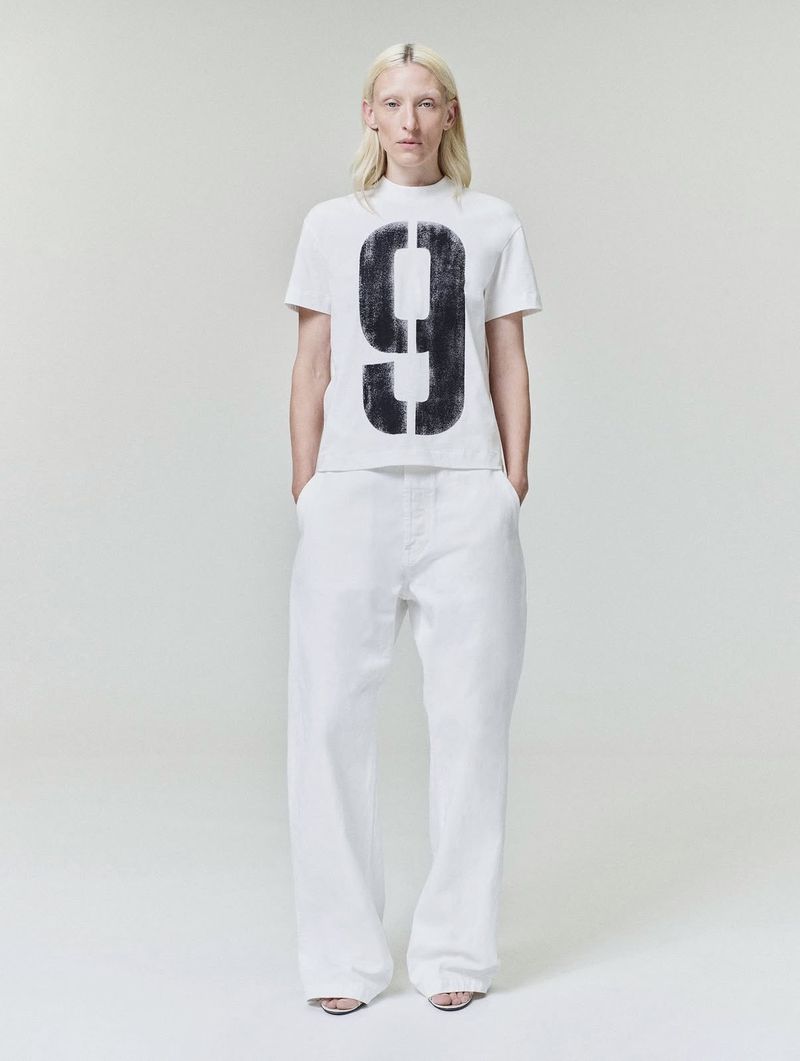 Model Maggie Maurer wearing a white t-shirt with a large number nine and white trousers against a seamless white background.