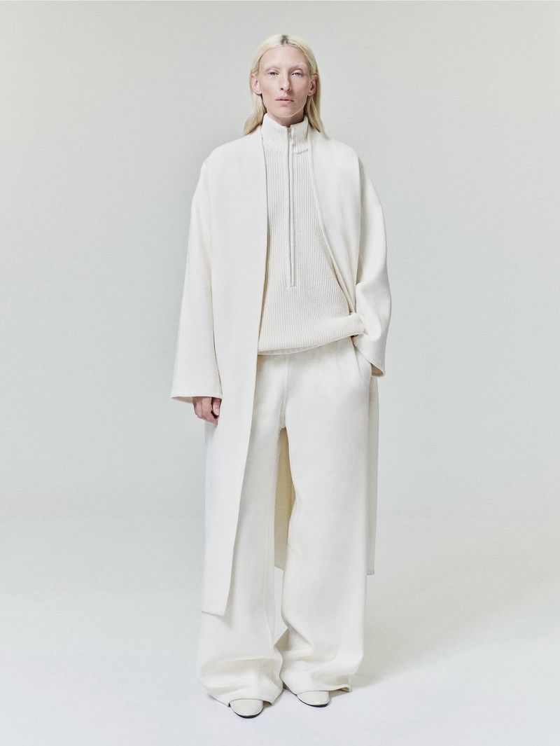 Model wearing a cream long coat, zip sweater, and wide trousers against a seamless white background.
