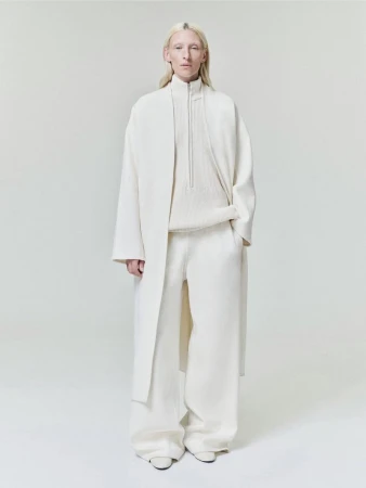 Model wearing a cream long coat, zip sweater, and wide trousers against a seamless white background.