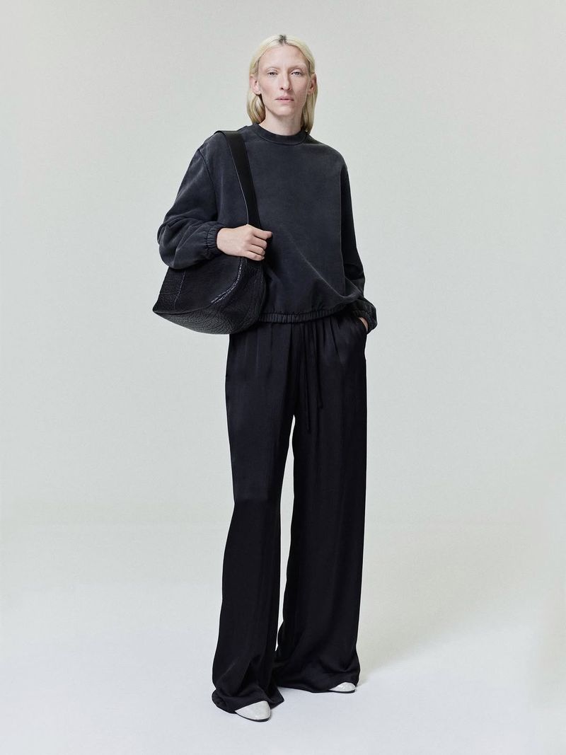 Model Maggie Maurer wearing a dark grey sweatshirt and wide black trousers carrying a shoulder bag against a neutral grey studio background