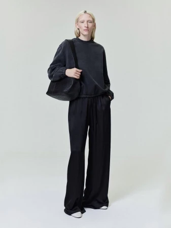 Model Maggie Maurer wearing a dark grey sweatshirt and wide black trousers carrying a shoulder bag against a neutral grey studio background