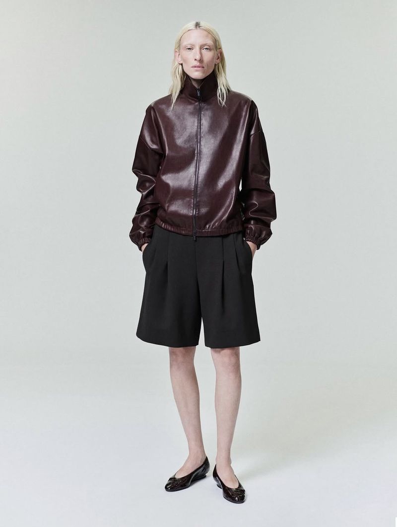 Model with blonde hair wearing a burgundy zip-up jacket and black shorts against a white backdrop.