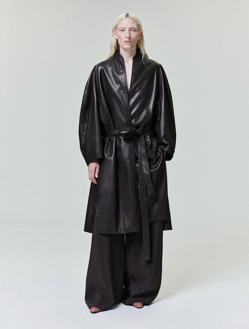 Model wearing a black leather coat with a belted waist against a neutral background
