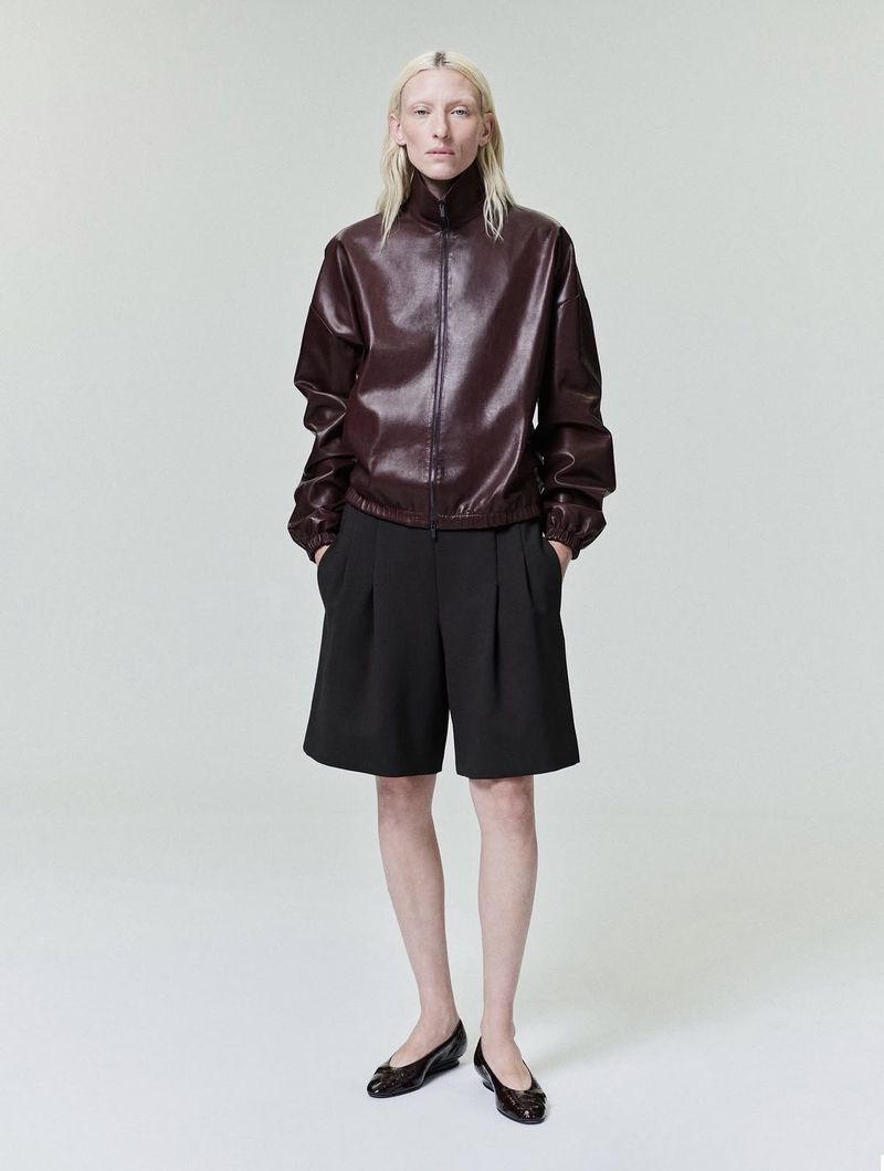 Model Maggie Maurer wearing a dark burgundy leather jacket and black pleated shorts against a seamless grey backdrop