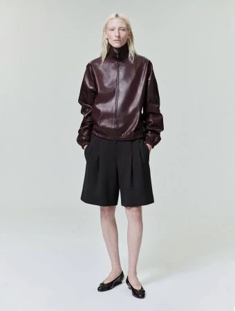 Model Maggie Maurer wearing a dark burgundy leather jacket and black pleated shorts against a seamless grey backdrop