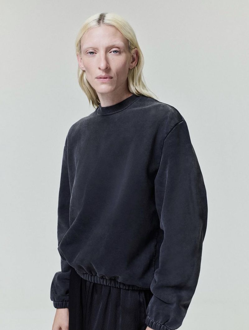 Model with long blonde hair wearing a dark grey crewneck sweatshirt against a plain grey studio background.