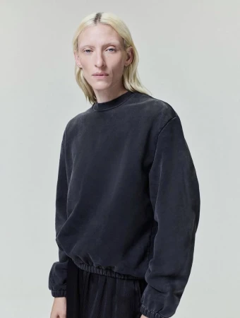 Model with long blonde hair wearing a dark grey crewneck sweatshirt against a plain grey studio background.