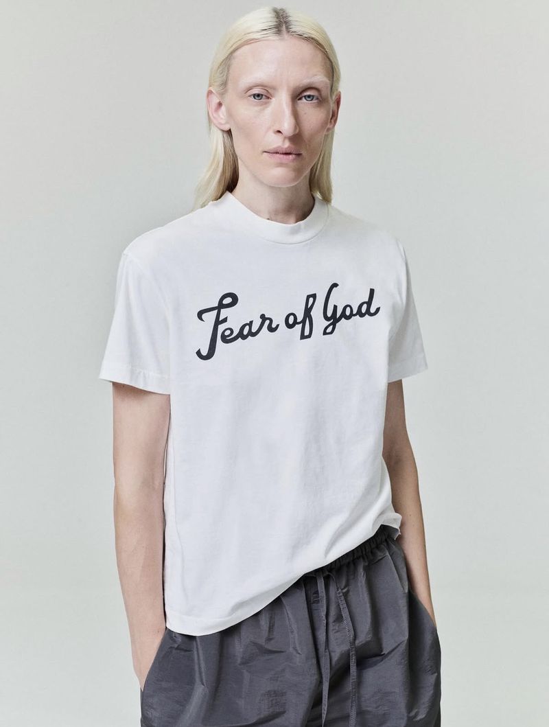 Model Maggie Maurer wearing a white graphic t-shirt and grey trousers against a light grey seamless background