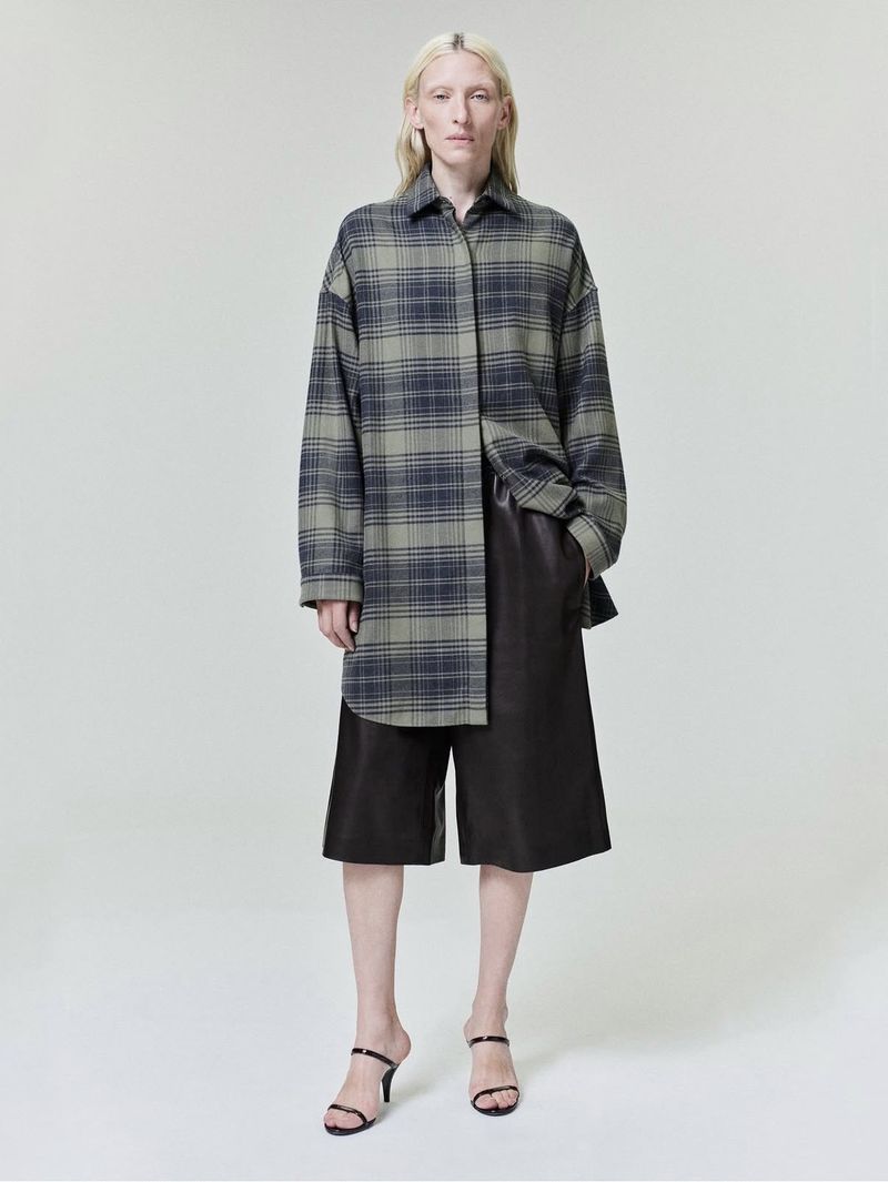 Model wearing an oversized plaid shirt and dark trousers against a neutral grey studio background.