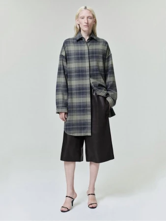 Model wearing an oversized plaid shirt and dark trousers against a neutral grey studio background.