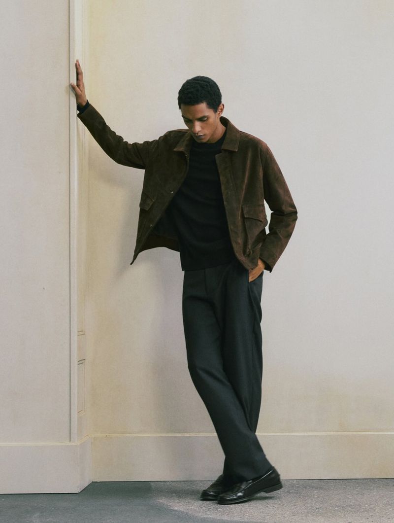 Model leaning against a wall wearing a brown suede jacket, black shirt, and dark trousers.