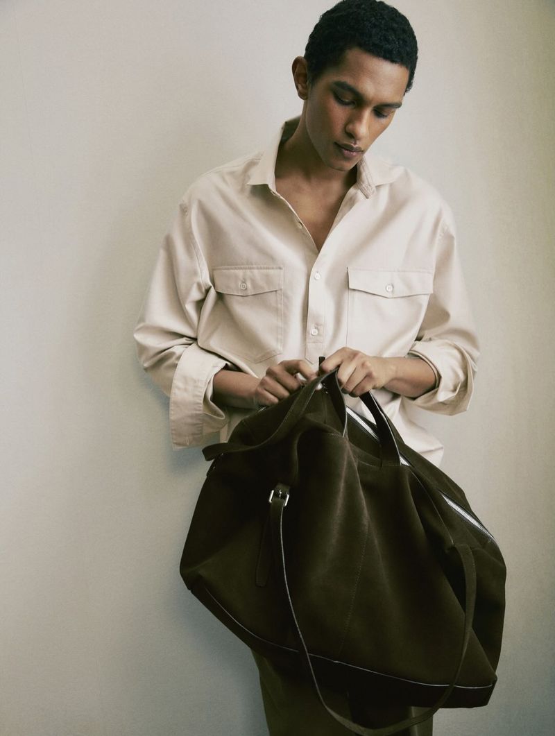 Man wearing a cream button-down shirt holding a dark leather duffel bag against a plain wall for Massimo Dutti