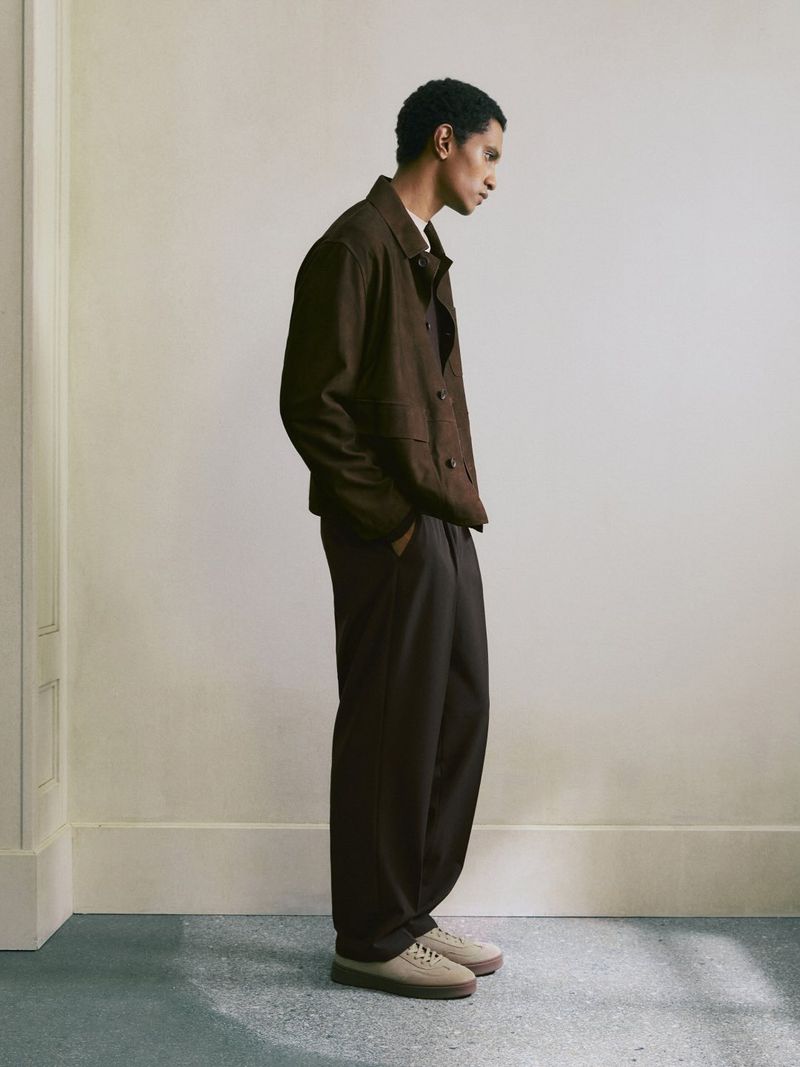 Profile view of a man standing against a plain wall wearing a brown jacket, dark trousers, and light sneakers.