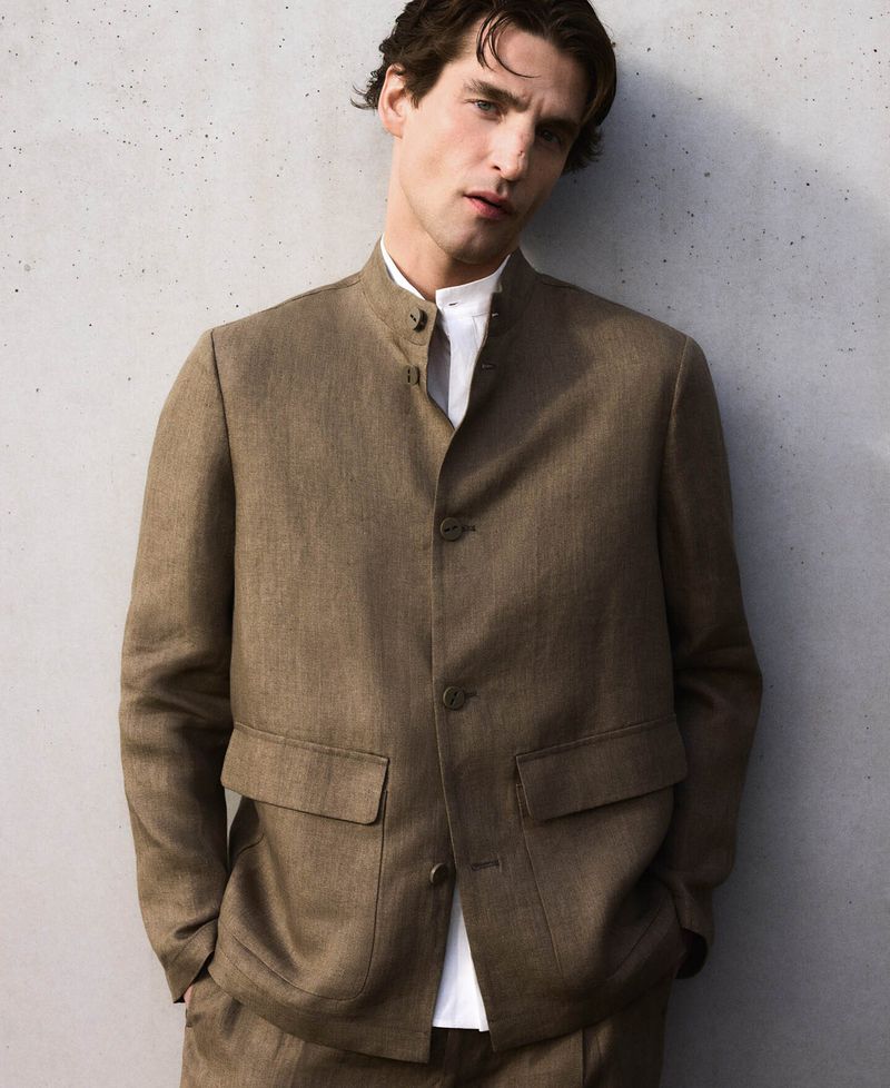 Man wearing a brown buttoned jacket and white shirt posing against a textured concrete wall.