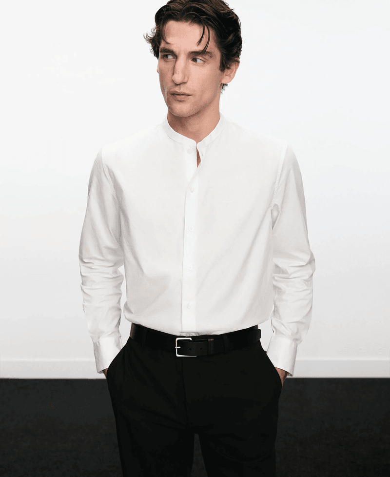 Model wearing a white mandarin-collar shirt and black trousers with hands in pockets against a plain white background.