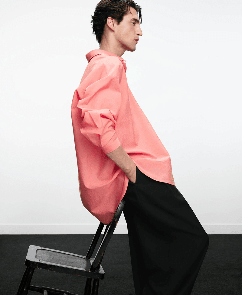 Model in a coral pink oversized shirt leaning back against a black chair against a white wall.