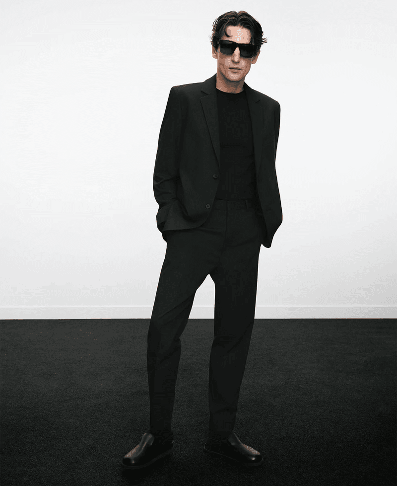 Man wearing a black suit and sunglasses standing against a plain white background for Adolfo Dominguez.
