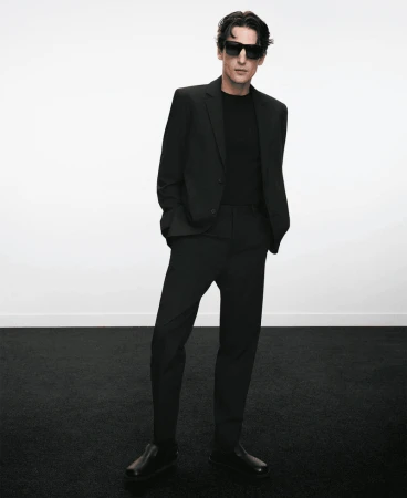 Man wearing a black suit and sunglasses standing against a plain white background for Adolfo Dominguez.