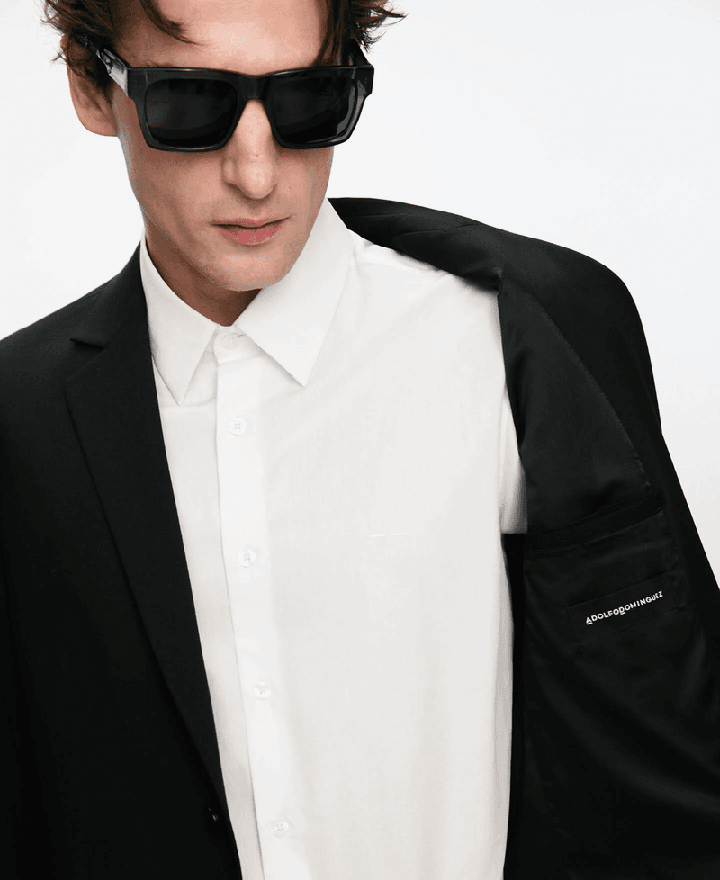 Man wearing dark sunglasses, a crisp white button-down shirt, and a black blazer against a solid white background.