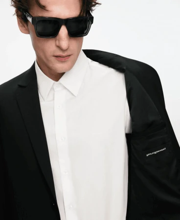 Man wearing dark sunglasses, a crisp white button-down shirt, and a black blazer against a solid white background.