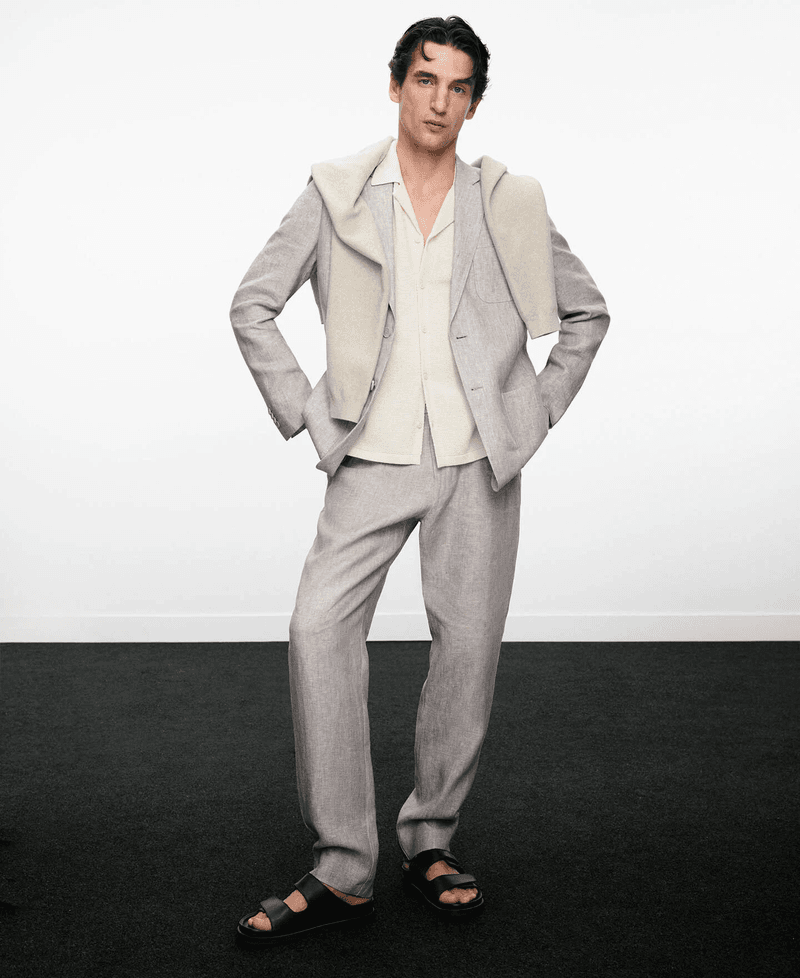 Full-length portrait of a male model wearing a grey suit, light shirt, and sweater draped over shoulders against a white wall.