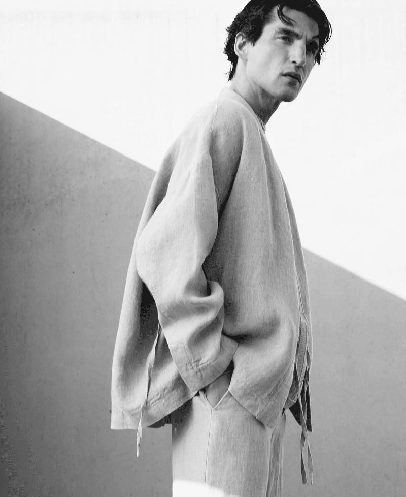 Monochrome fashion portrait of a man wearing a loose linen jacket and trousers against a minimalist architectural wall.