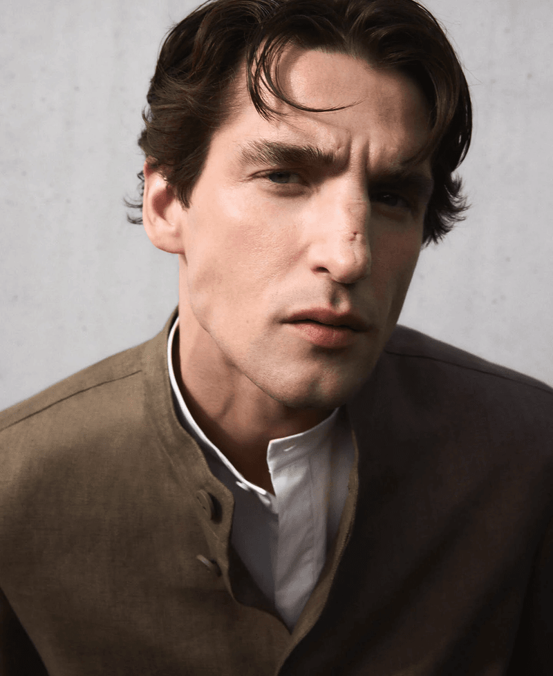 Portrait of a male model in a brown jacket and white collar shirt against a neutral textured background.