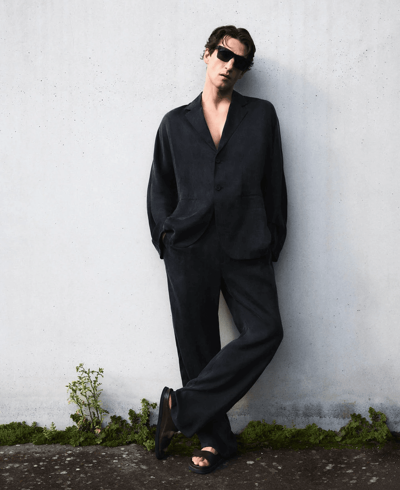 A man wearing a black suit and sunglasses leans against a concrete wall with moss, showcasing a fashion-forward, relaxed pose.