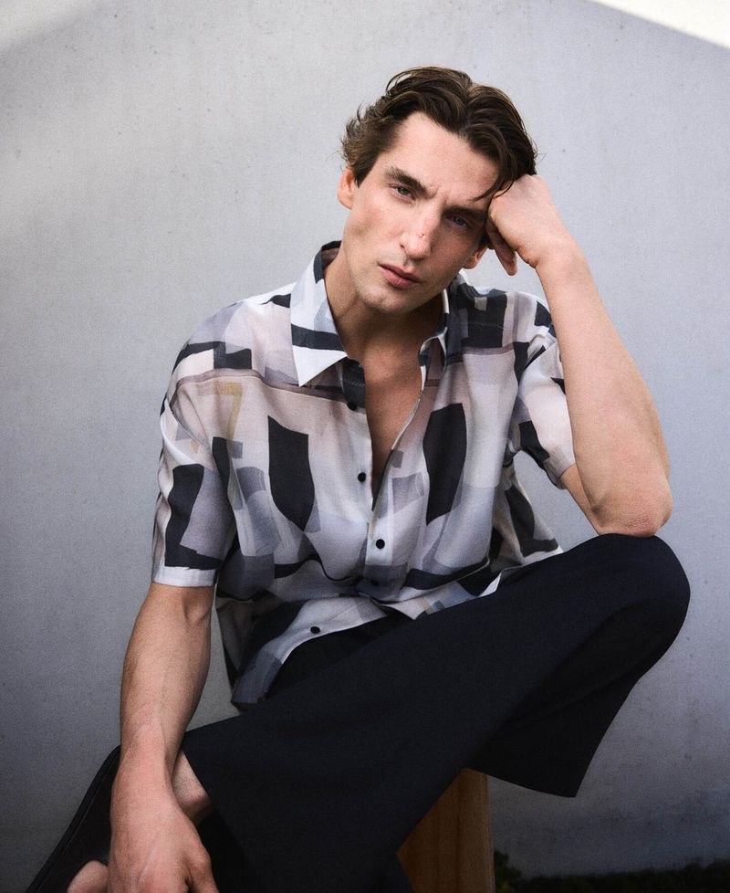 Model Anatol Modzelewski wearing a patterned short-sleeve shirt and dark trousers against a neutral wall for Adolfo Dominguez.