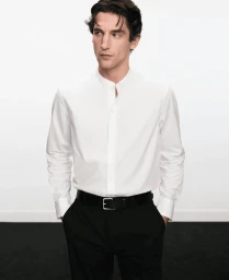 Model wearing a white mandarin-collar shirt and black trousers with hands in pockets against a plain white background.
