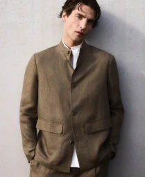Man wearing a brown buttoned jacket and white shirt posing against a textured concrete wall.