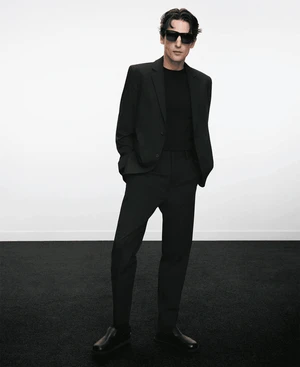 Man wearing a black suit and sunglasses standing against a plain white background for Adolfo Dominguez.