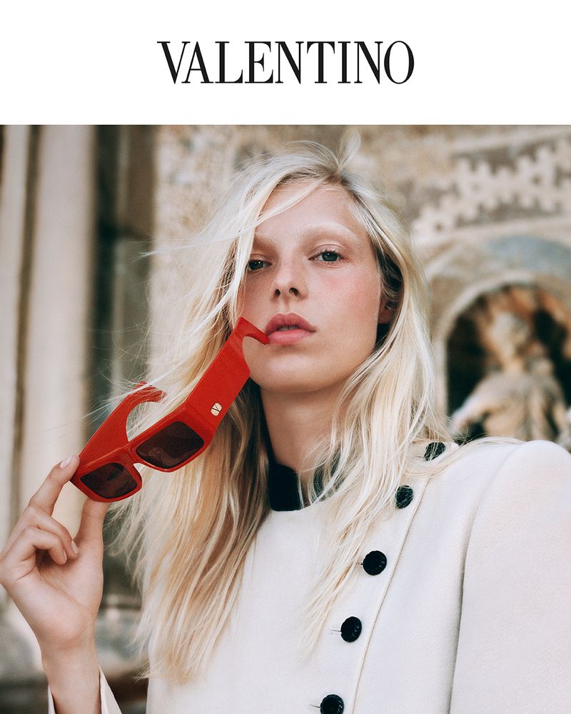 Model holding bold red sunglasses wearing an off-white buttoned jacket with a classical architectural background.