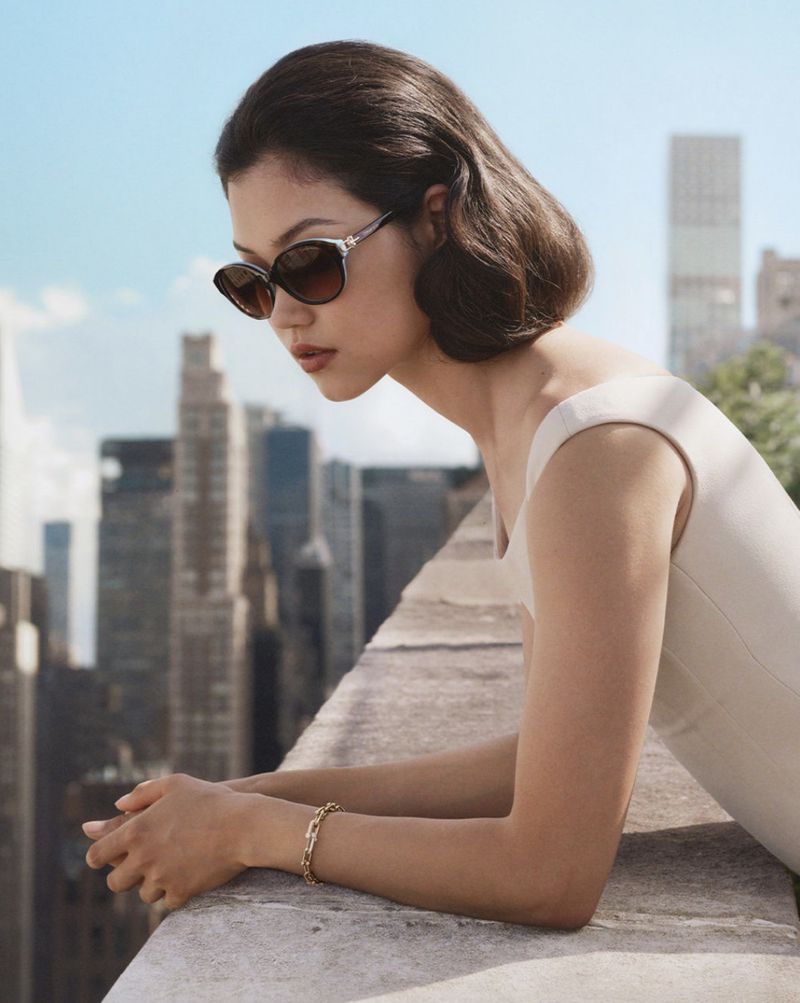 A stylish woman with short brown hair wears sunglasses and a light-colored dress while leaning on a stone ledge with a city skyline background.