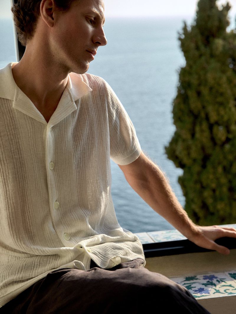 Man in white shirt sits by a window overlooking the sea. Green foliage partially obscures the view in soft, natural light.