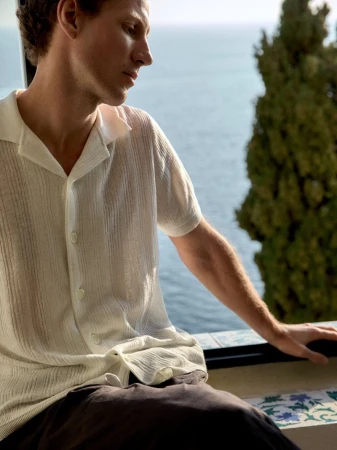 Man in white shirt sits by a window overlooking the sea. Green foliage partially obscures the view in soft, natural light.
