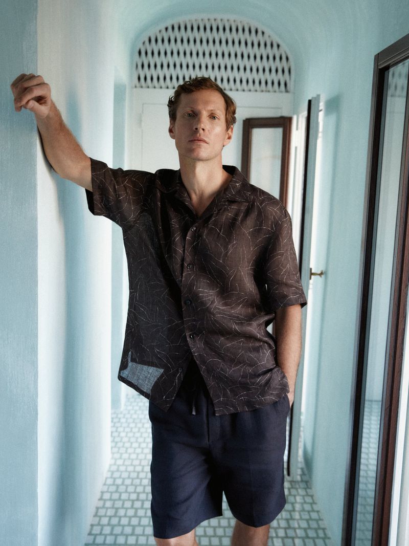 Man wearing a brown patterned short-sleeve shirt and dark shorts standing in a light blue hallway.