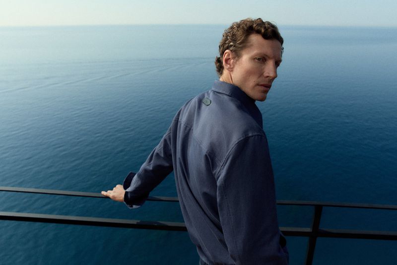 Man wearing a blue jacket leaning on a black railing with the ocean visible in the background