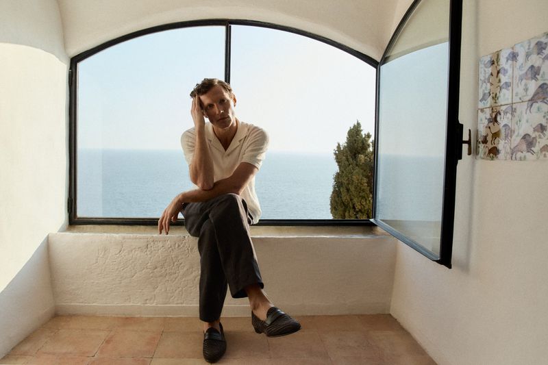 Man seated on a window ledge wearing a cream polo shirt and dark trousers with ocean view in background