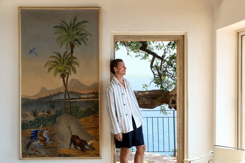 Man wearing a striped jacket leaning against a doorway next to a large framed tropical landscape painting