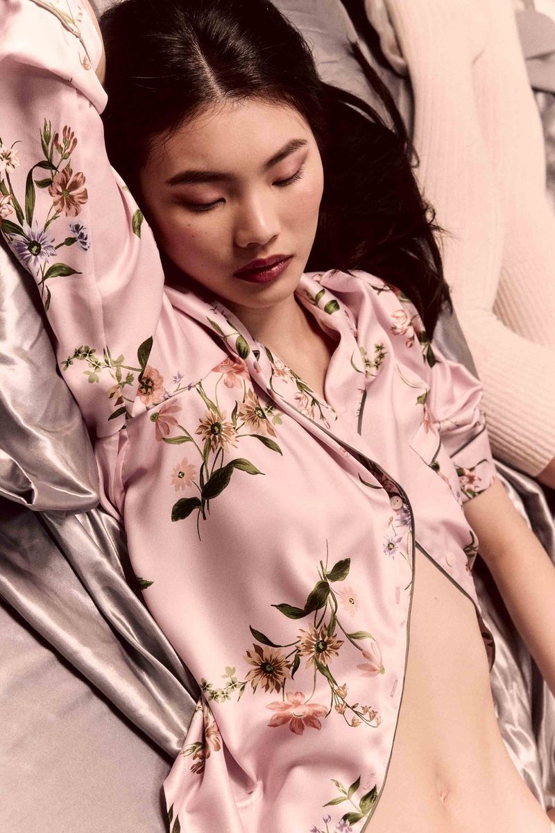 Model wearing pink floral silk pajama top resting eyes closed on satin sheets