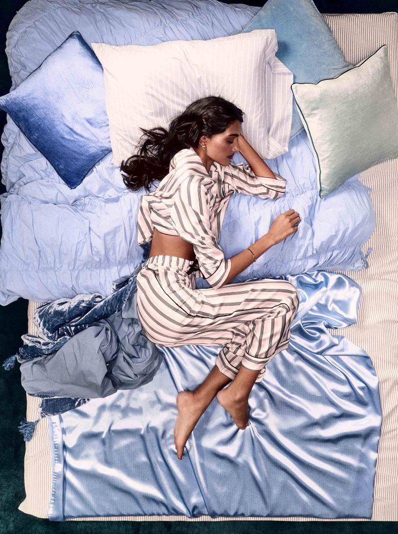 Woman in striped pajamas sleeps peacefully on a blue-toned bed covered with pillows and a throw, captured in a high-angle shot.
