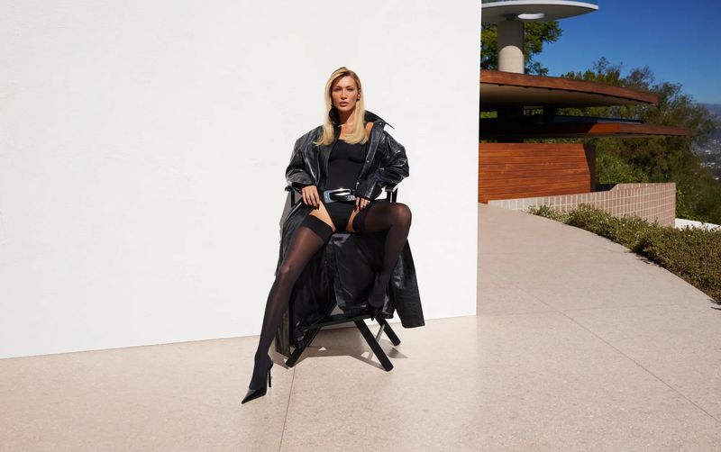Bella Hadid in a black leather coat and bodysuit sitting on a director chair against a white wall for Revolve S/S 2026