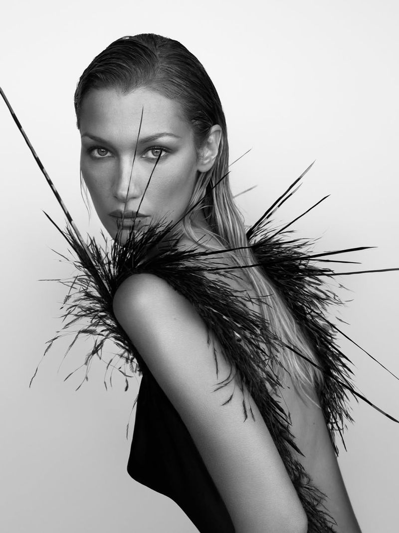 Monochrome portrait of model Bella Hadid wearing a black feathered garment against a plain white background for Revolve.