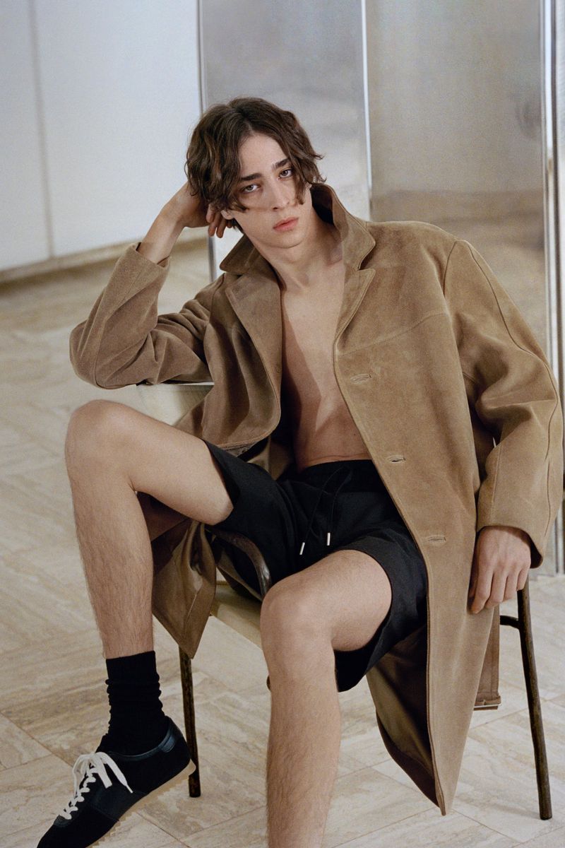 Young male model seated on a chair wearing an open camel coat, black shorts, and sneakers against a minimalist indoor background.